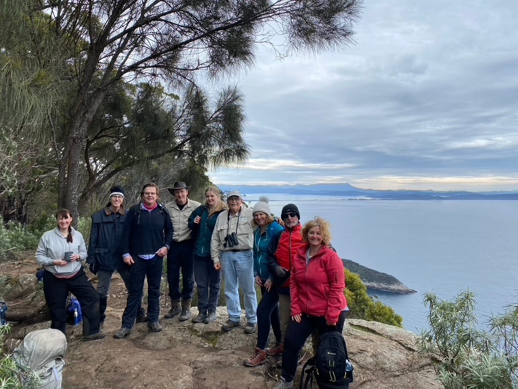 Bruny Island-elementor-io-optimized | Park Trek Group of hikers posing on a rocky cliff with trees, overlooking a calm sea and distant mountains.
