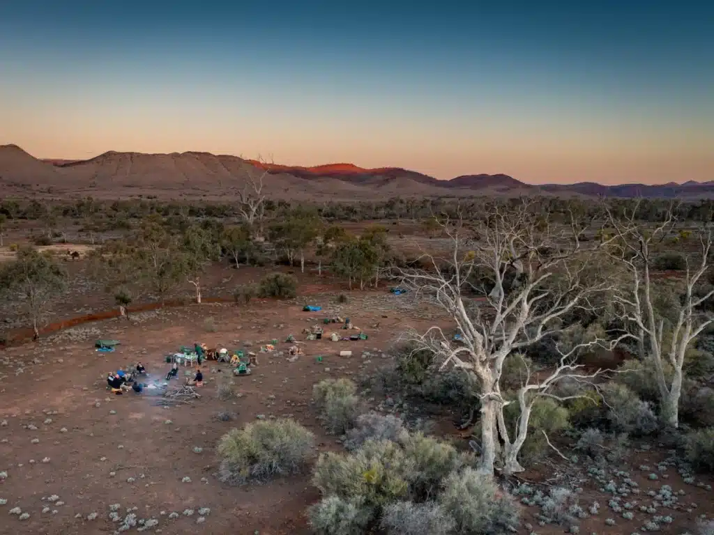 Desert campsite at sunset with a campfire circle, tents, and people gathering among sparse trees under a clear sky.