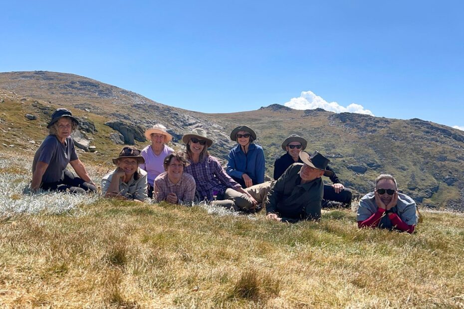 Ten hikers lying on a sunlit alpine meadow, smiling with a rocky hill and blue sky behind them.