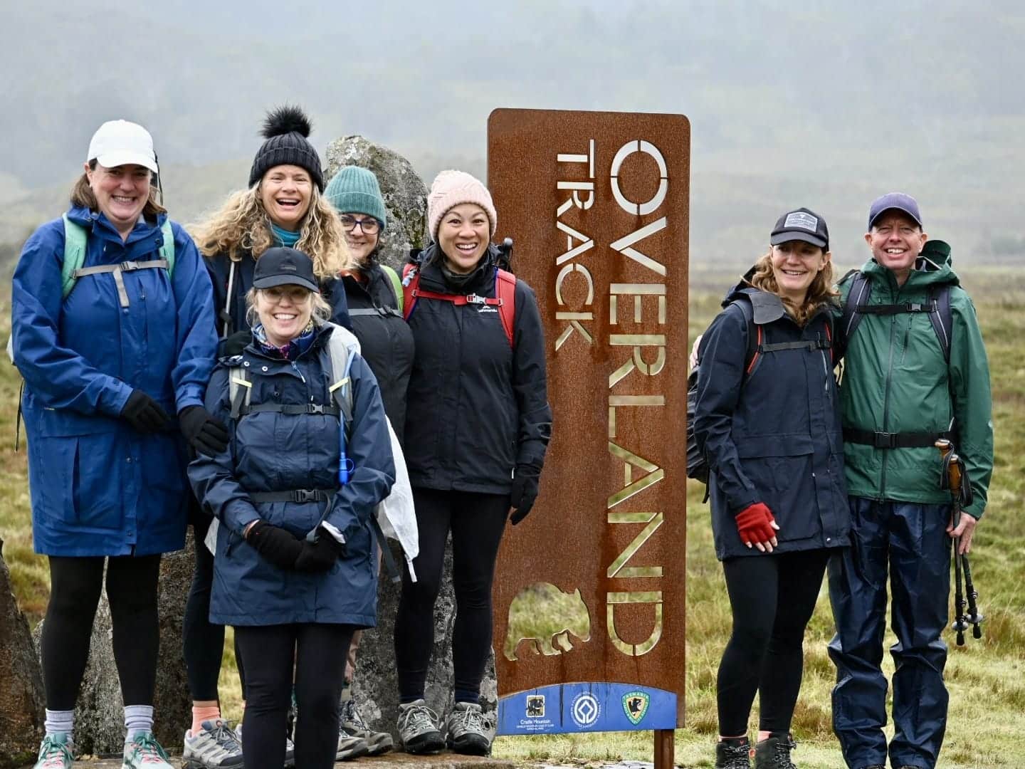 Overland Track-elementor-io-optimized | Park Trek Group of seven hikers in rain jackets and backpacks posing beside a rust-colored'Overland Track' sign in a misty landscape.