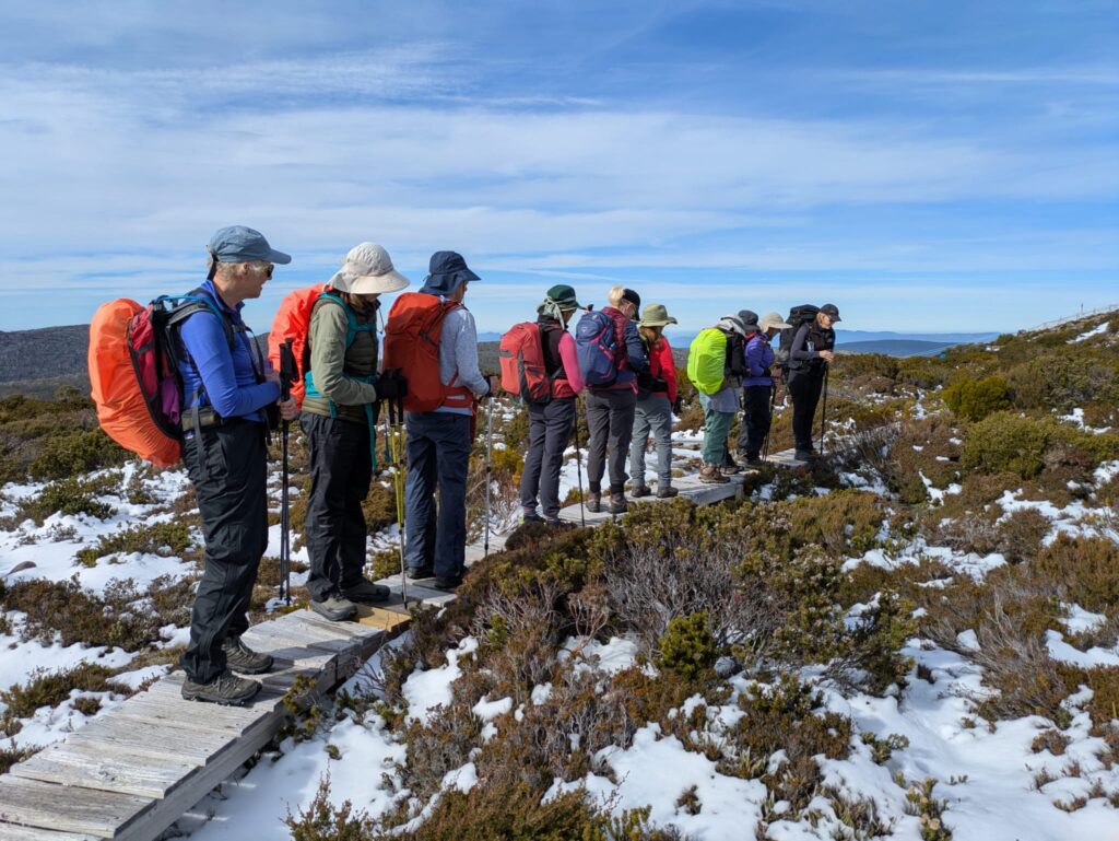 Group of hikers standing on a wooden boardwalk in a snowy, rocky landscape, wearing backpacks and hats.