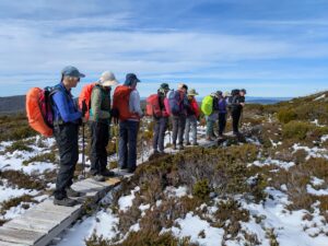 Group of hikers standing on a wooden boardwalk in a snowy, rocky landscape, wearing backpacks and hats.