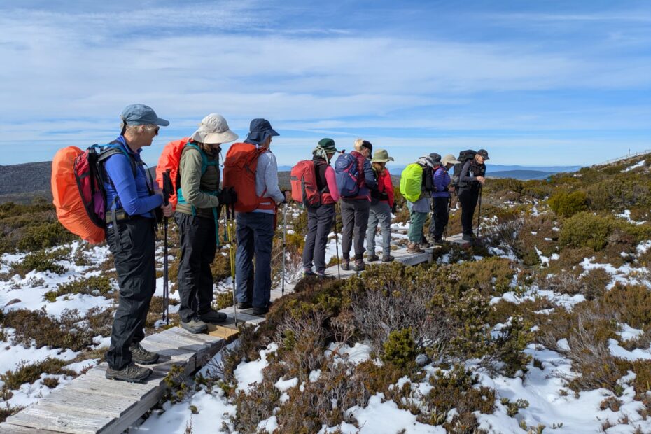 Group of hikers standing on a wooden boardwalk in a snowy, rocky landscape, wearing backpacks and hats.